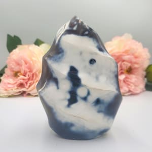 May include: A polished stone sculpture with a unique, organic shape. The stone features a blend of white and dark blue hues, creating a marbled effect. The sculpture is set against a soft, blurred background with pink flowers.