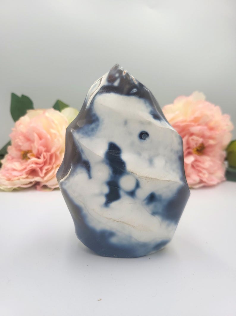 May include: A polished stone sculpture with a unique, organic shape. The stone features a blend of white and dark blue hues, creating a marbled effect. The sculpture is set against a soft, blurred background with pink flowers.