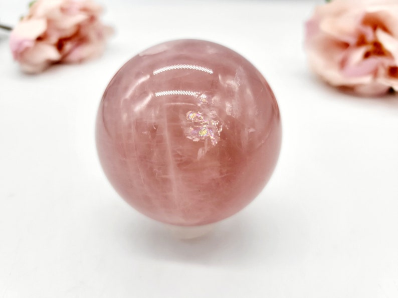 May include: A large, polished rose quartz sphere with a pink hue and iridescent inclusions. The sphere is smooth and reflective, with visible striations in the stone.