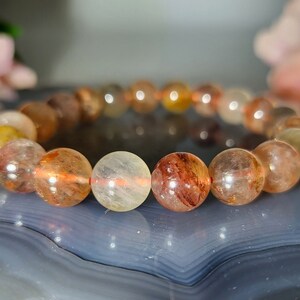 May include: A beaded bracelet featuring translucent, spherical beads in shades of yellow, orange, and brown. The beads are strung together, creating a circular shape. The bracelet is displayed on a blue and gray surface.