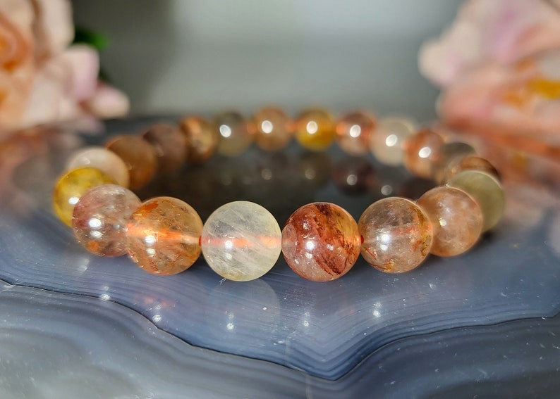 May include: A beaded bracelet featuring translucent, spherical beads in shades of yellow, orange, and brown. The beads are strung together, creating a circular shape. The bracelet is displayed on a blue and gray surface.