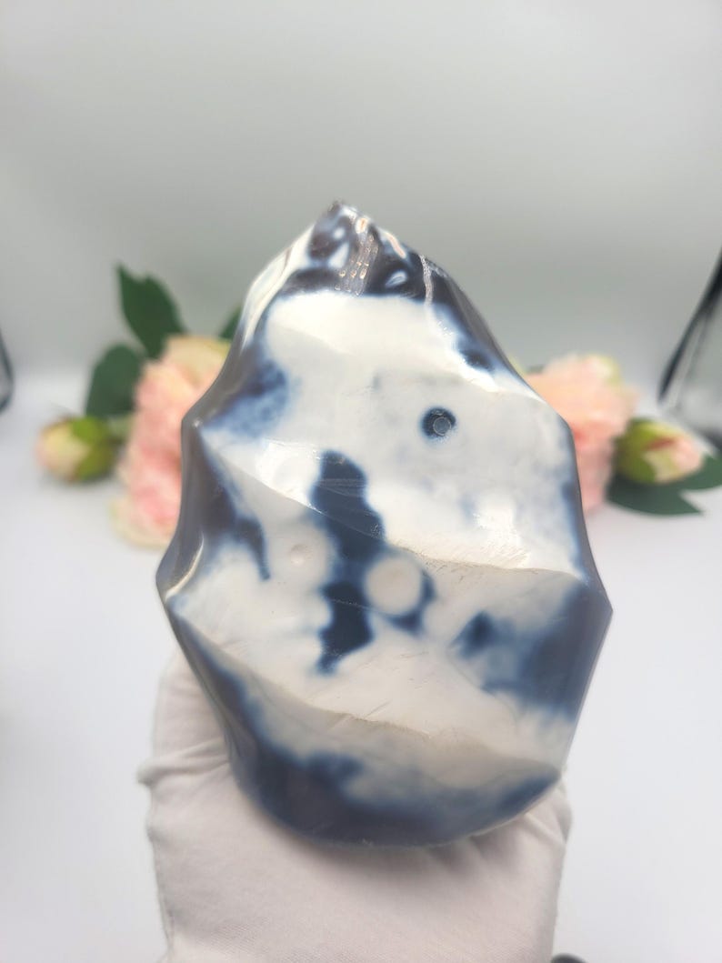 May include: A polished, freeform stone with a marbled appearance. The stone features a mix of white and deep blue hues, creating a unique pattern. The shape is irregular, with rounded edges and a smooth, glossy surface. The stone is held by a gloved hand.