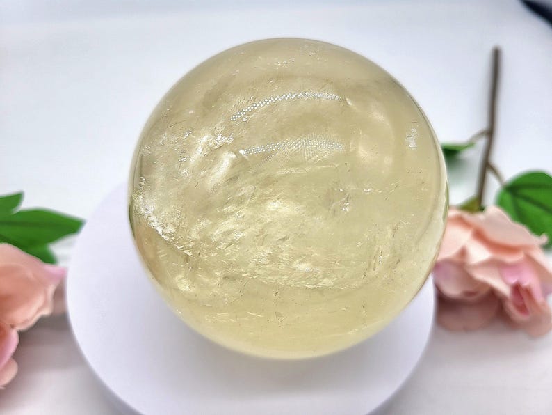 May include: A large, smooth, yellow citrine sphere on a white stand. The sphere has a translucent, glassy appearance with subtle internal striations.