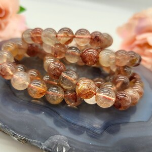 May include: Two bracelets made of clear, round beads with reddish-brown and yellow inclusions. The bracelets are resting on a gray and blue marbled surface, with a blurred floral background.