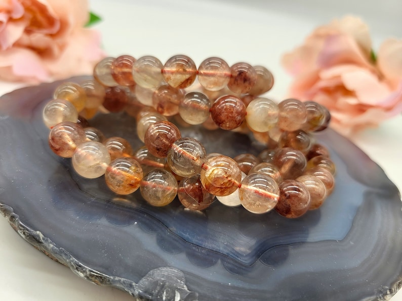 May include: Two bracelets made of clear, round beads with reddish-brown and yellow inclusions. The bracelets are resting on a gray and blue marbled surface, with a blurred floral background.