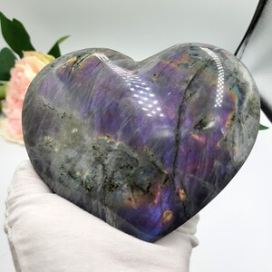 May include: A heart-shaped labradorite stone with a polished surface. The stone displays a mix of gray, purple, and iridescent colors. The heart is held by a gloved hand, showcasing the stone's size and shape. The stone is a decorative item.