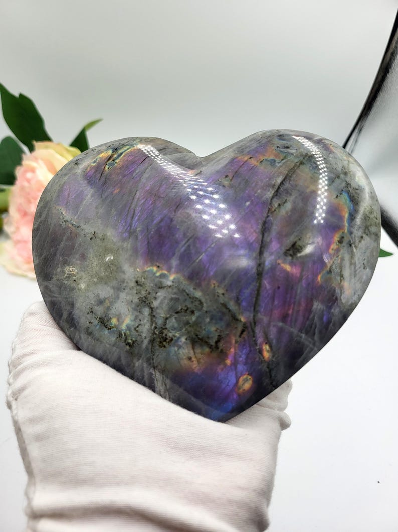 May include: A heart-shaped labradorite stone with a polished surface. The stone displays a mix of gray, purple, and iridescent colors. The heart is held by a gloved hand, showcasing the stone's size and shape. The stone is a decorative item.