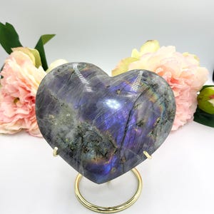 May include: A heart-shaped labradorite stone with iridescent purple and blue hues, displayed on a gold-colored metal stand. The stone is polished and smooth, with natural patterns. Pink and yellow flowers are in the background.
