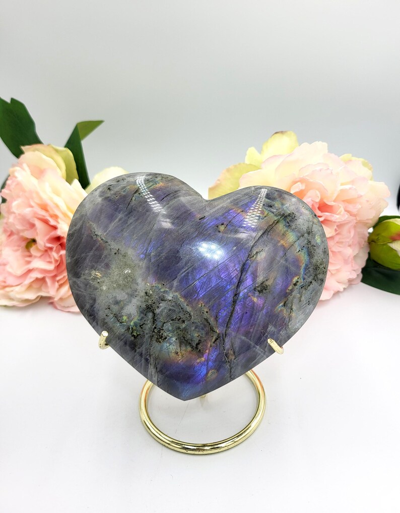 May include: A heart-shaped labradorite stone with iridescent purple and blue hues, displayed on a gold-colored metal stand. The stone is polished and smooth, with natural patterns. Pink and yellow flowers are in the background.