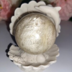 May include: A pearlescent sphere rests in a white seashell-shaped holder, with a small dolphin figure on the side. The sphere has a shimmering, textured surface. The background features soft pink and green floral elements.