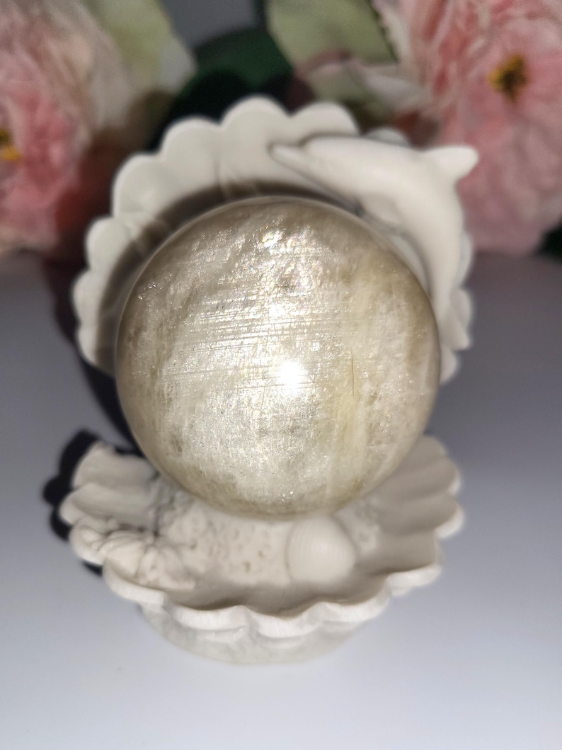 May include: A pearlescent sphere rests in a white seashell-shaped holder, with a small dolphin figure on the side. The sphere has a shimmering, textured surface. The background features soft pink and green floral elements.