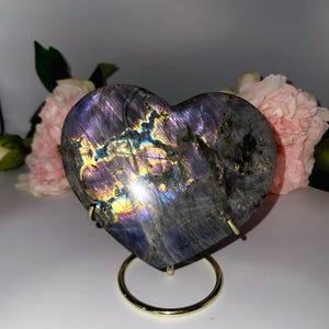 May include: A heart-shaped labradorite stone with iridescent colors of purple, blue, and gold. The stone is displayed on a gold-colored metal stand. Pink flowers are in the background.