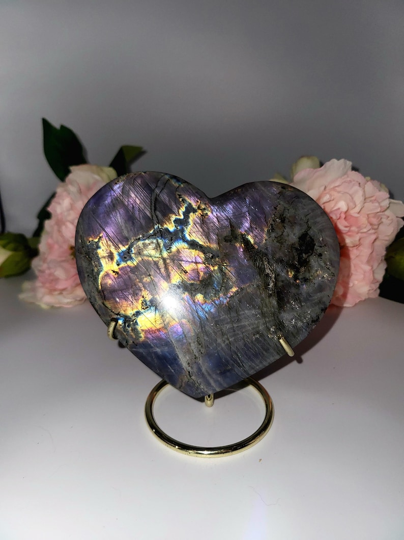 May include: A heart-shaped labradorite stone with iridescent colors of purple, blue, and gold. The stone is displayed on a gold-colored metal stand. Pink flowers are in the background.