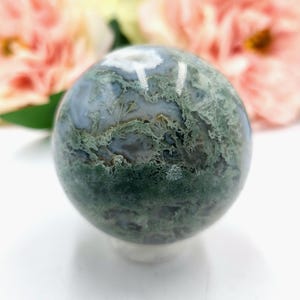 K&ouml;nnte beinhalten: Eine polierte Moosachat-Kugel, die eine Mischung aus gr&uuml;nen, wei&szlig;en und grauen Mustern zeigt. Der Edelsteinball wird auf einer wei&szlig;en Oberfl&auml;che pr&auml;sentiert, mit zarten rosa und gelben Blumen im Hintergrund.
