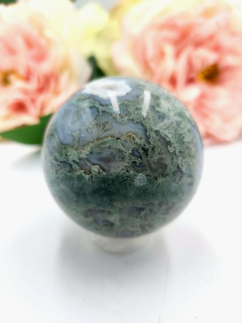 K&ouml;nnte beinhalten: Eine polierte Moosachat-Kugel, die eine Mischung aus gr&uuml;nen, wei&szlig;en und grauen Mustern zeigt. Der Edelsteinball wird auf einer wei&szlig;en Oberfl&auml;che pr&auml;sentiert, mit zarten rosa und gelben Blumen im Hintergrund.