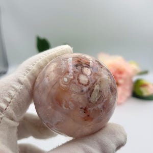 May include: A spherical gemstone, polished to a high sheen, displaying a mix of pink, cream, and brown tones. The orb-shaped crystal has a distinctive pattern of swirls and inclusions. A gloved hand holds the sphere. Pink flowers are in the background.