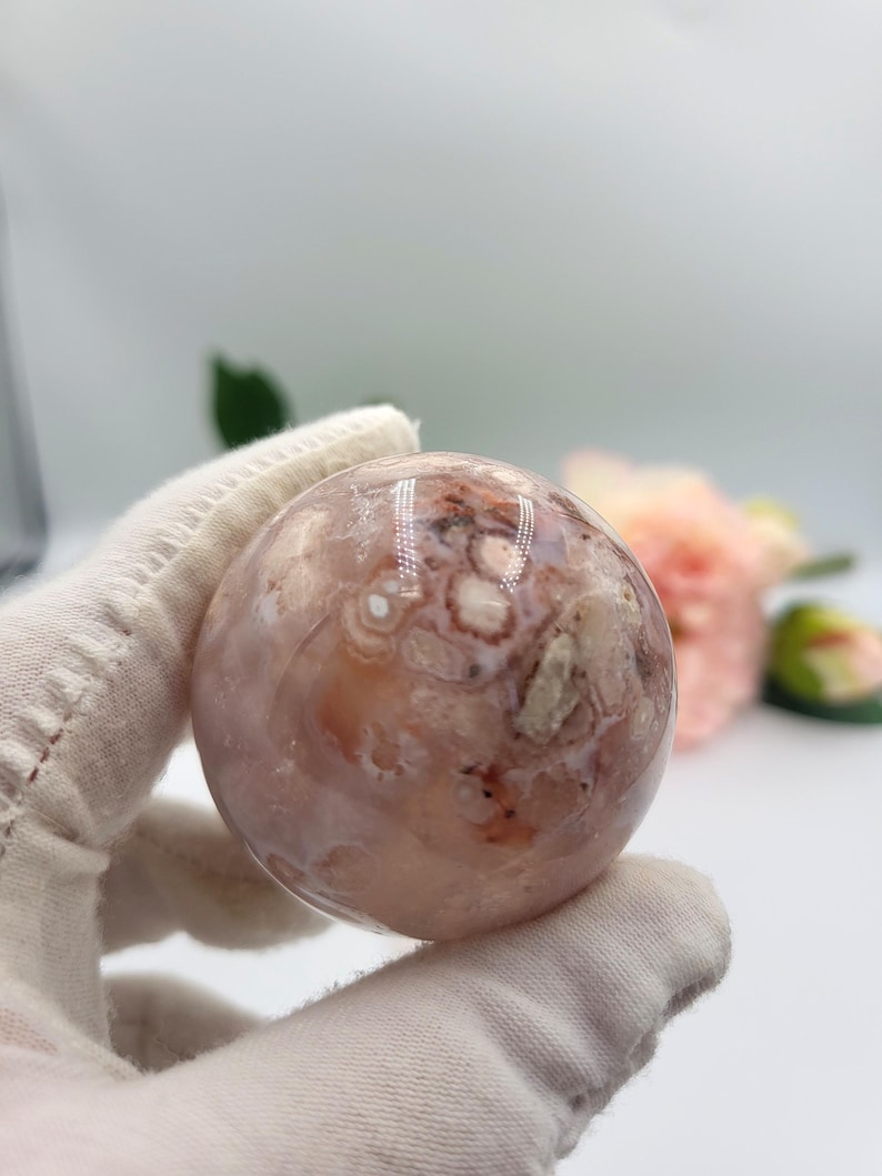 May include: A spherical gemstone, polished to a high sheen, displaying a mix of pink, cream, and brown tones. The orb-shaped crystal has a distinctive pattern of swirls and inclusions. A gloved hand holds the sphere. Pink flowers are in the background.