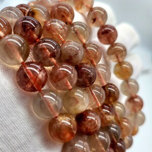 May include: A close-up of several strands of gemstone bead bracelets. The beads are round, translucent, and range in color from light amber to deep reddish-brown. Each bead has internal reddish inclusions. The bracelets are strung on a red cord.