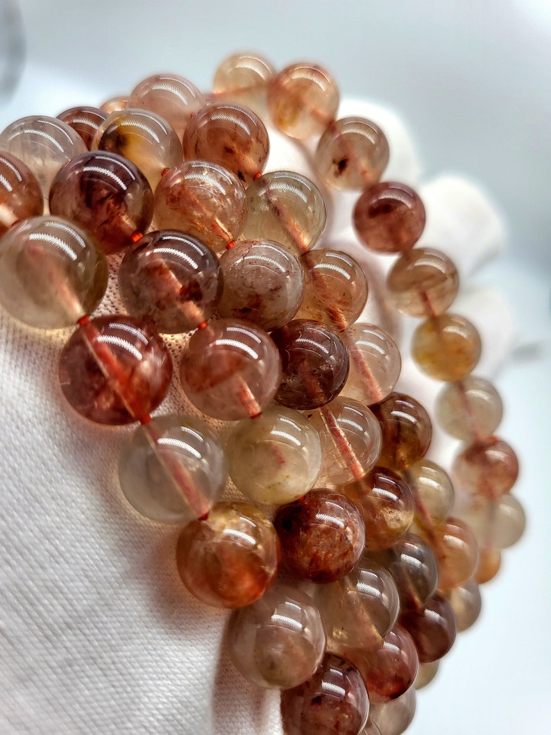 May include: A close-up of several strands of gemstone bead bracelets. The beads are round, translucent, and range in color from light amber to deep reddish-brown. Each bead has internal reddish inclusions. The bracelets are strung on a red cord.