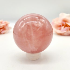 May include: A pink rose quartz sphere on a white stand. The sphere is smooth and polished, with a translucent appearance. The sphere is surrounded by a white background.