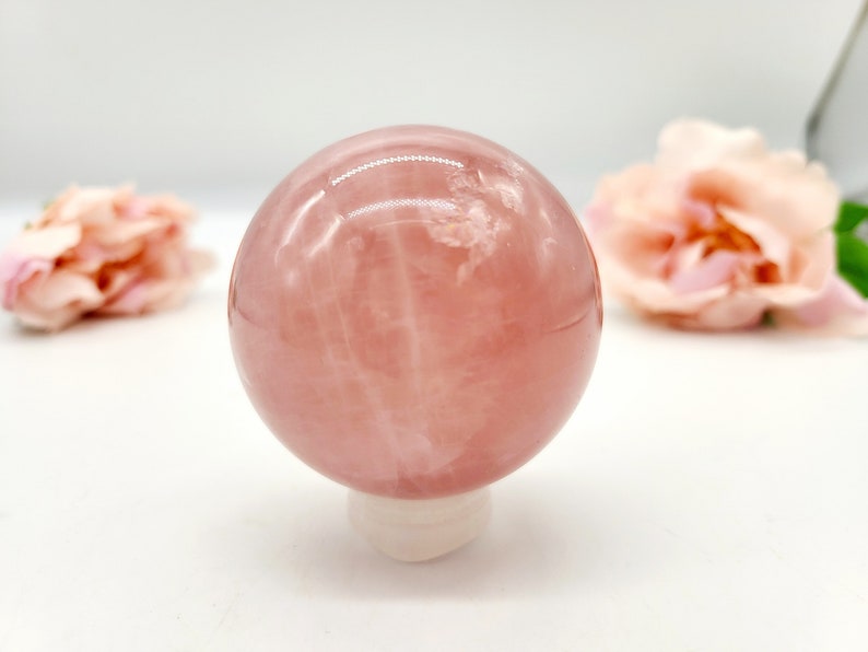 May include: A pink rose quartz sphere on a white stand. The sphere is smooth and polished, with a translucent appearance. The sphere is surrounded by a white background.