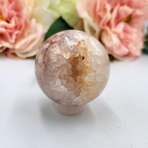 May include: A spherical, pale pink geode crystal ball with a hollowed-out centre filled with sparkling, light orange crystals. The crystal ball sits on a small, round base. Soft pink flowers are in the background.