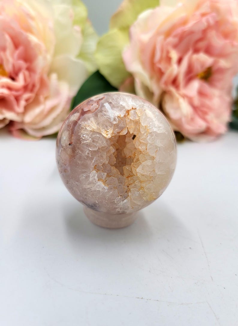 May include: A spherical, pale pink geode crystal ball with a hollowed-out centre filled with sparkling, light orange crystals. The crystal ball sits on a small, round base. Soft pink flowers are in the background.