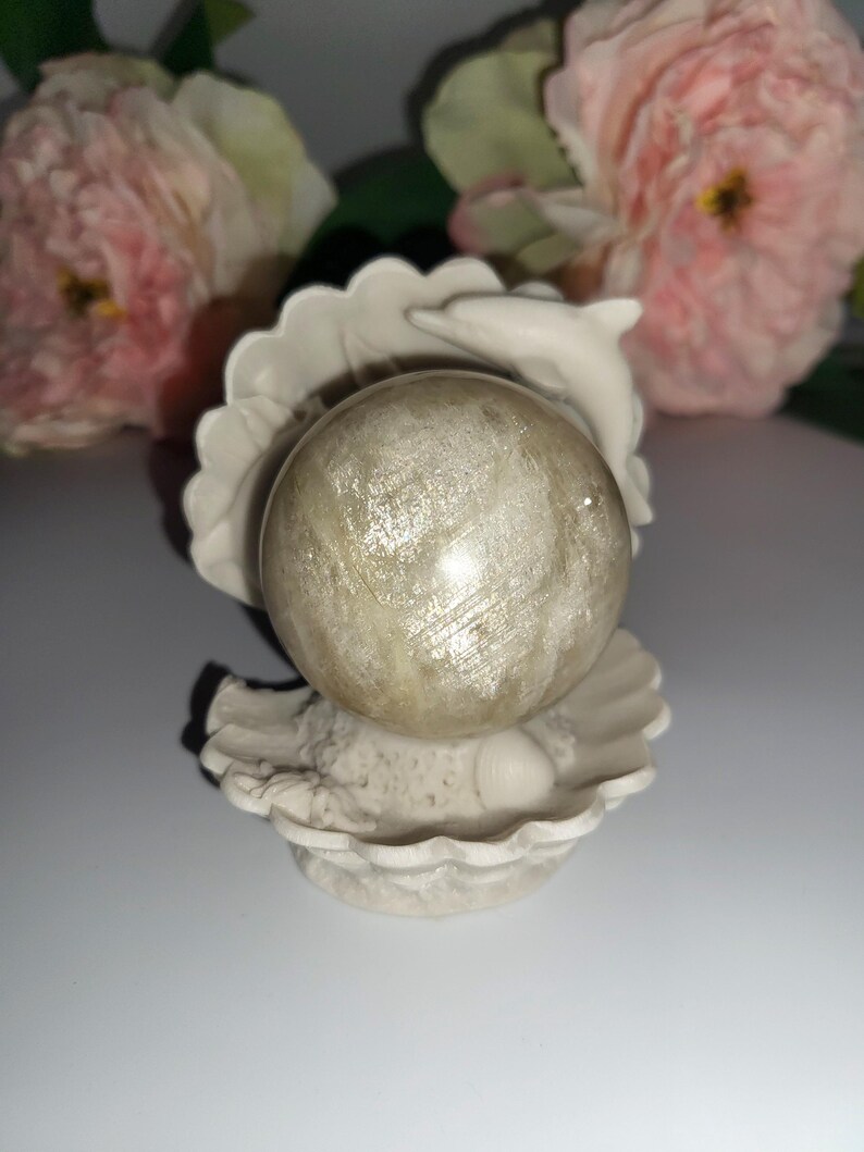 May include: A decorative, off-white seashell-shaped holder with a sphere. The sphere is a light, pearlescent color with subtle striations. The holder has a dolphin design and is set against a blurred floral background.