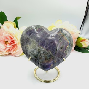May include: A heart-shaped, polished stone with a mix of purple, gray, and iridescent colors. The stone is displayed on a gold-colored metal stand with a circular base. The background features soft pink and yellow flowers.