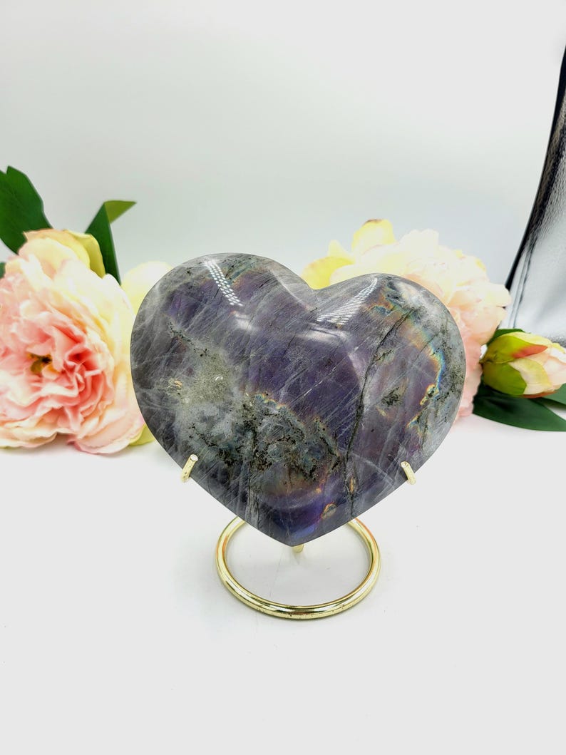 May include: A heart-shaped, polished stone with a mix of purple, gray, and iridescent colors. The stone is displayed on a gold-colored metal stand with a circular base. The background features soft pink and yellow flowers.
