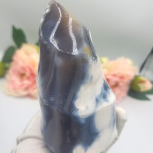 May include: A polished, natural stone sculpture with a smooth, rounded shape. The stone features a blend of colors, including white, blue, and brown, creating a marbled effect. The sculpture is held by a gloved hand, with a soft background of flowers.