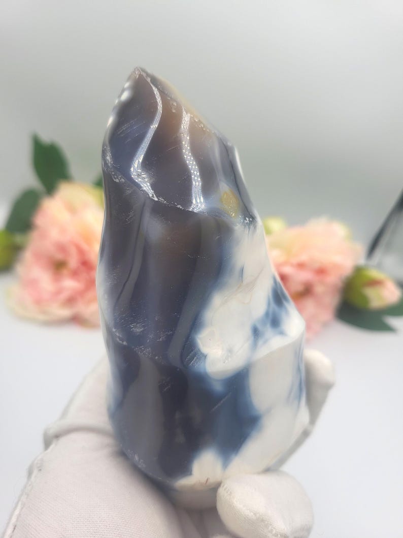 May include: A polished, natural stone sculpture with a smooth, rounded shape. The stone features a blend of colors, including white, blue, and brown, creating a marbled effect. The sculpture is held by a gloved hand, with a soft background of flowers.