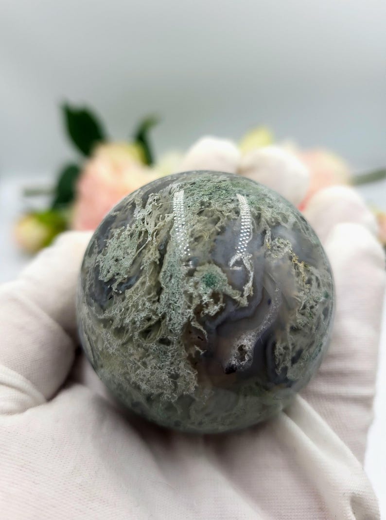 K&ouml;nnte beinhalten: Ein polierter, kugelf&ouml;rmiger Edelstein mit einem moosgr&uuml;nen und grauen Muster. Die Kugel wird in einer behandschuhten Hand gehalten, mit unscharfen Blumen im Hintergrund. Die Oberfl&auml;che des Steins hat ein nat&uuml;rliches, organisches Aussehen.