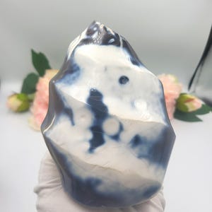 May include: A polished, freeform stone with a marbled appearance. The stone features a mix of white and dark blue hues, creating a unique pattern. The shape is irregular, with a smooth, glossy surface. The stone is held by a gloved hand, suggesting it is a decorative item.