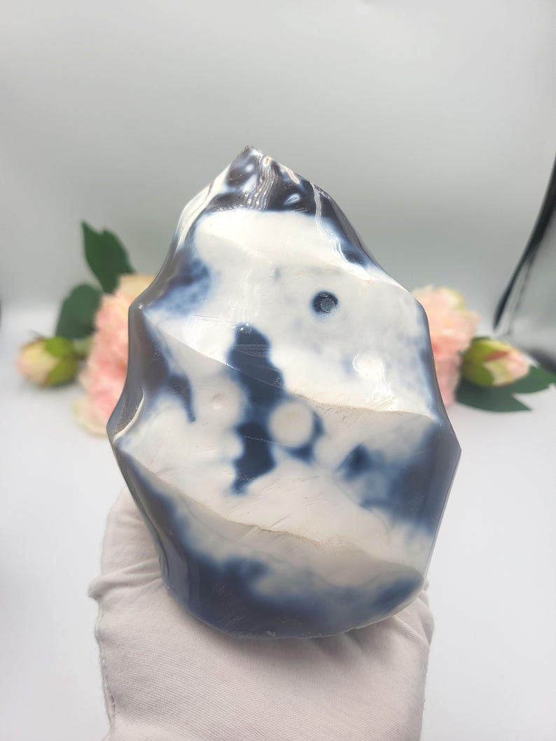 May include: A polished, freeform stone with a marbled appearance. The stone features a mix of white and dark blue hues, creating a unique pattern. The shape is irregular, with a smooth, glossy surface. The stone is held by a gloved hand, suggesting it is a decorative item.