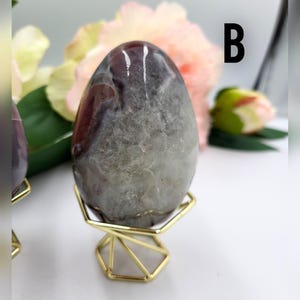 May include: A polished, egg-shaped stone with a mix of gray, brown, and white hues. The stone is displayed on a small, gold-colored geometric stand. The letter "B" is visible in the background.