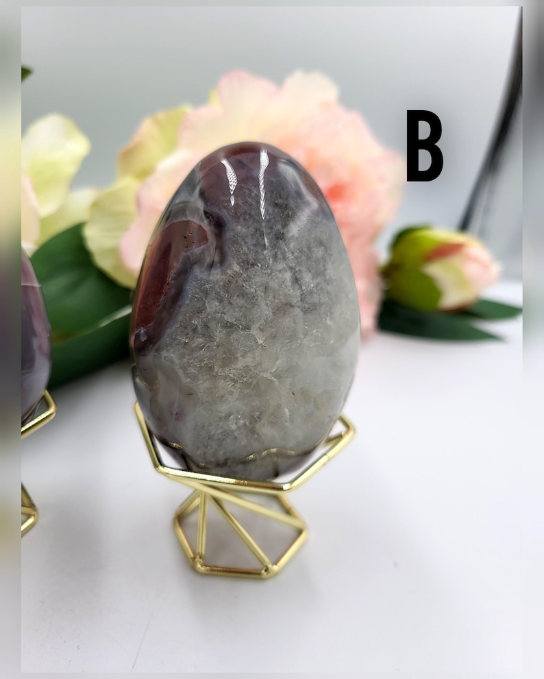 May include: A polished, egg-shaped stone with a mix of gray, brown, and white hues. The stone is displayed on a small, gold-colored geometric stand. The letter "B" is visible in the background.