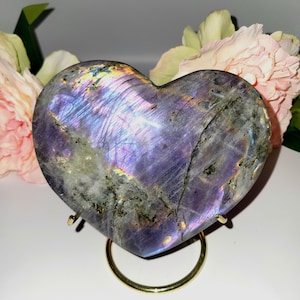 May include: A heart-shaped labradorite stone with iridescent purple, blue, and gold hues. The polished stone sits on a gold-colored metal stand, with pink and cream-colored flowers in the background. The stone's surface has a natural, textured appearance.