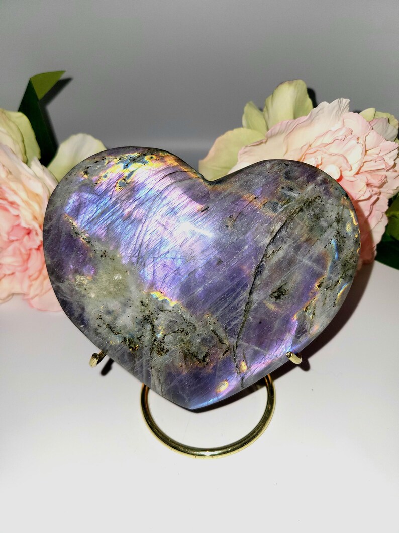 May include: A heart-shaped labradorite stone with iridescent purple, blue, and gold hues. The polished stone sits on a gold-colored metal stand, with pink and cream-colored flowers in the background. The stone's surface has a natural, textured appearance.