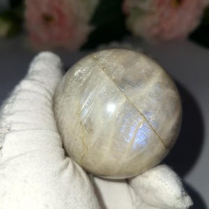 May include: A round, iridescent moonstone sphere. The sphere has a pearly white base color with flashes of blue and a visible crack. The sphere is held by a gloved hand, with soft pink flowers in the background.