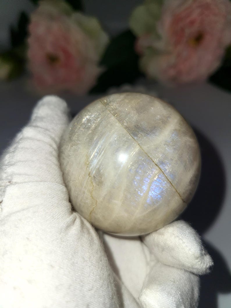 May include: A round, iridescent moonstone sphere. The sphere has a pearly white base color with flashes of blue and a visible crack. The sphere is held by a gloved hand, with soft pink flowers in the background.
