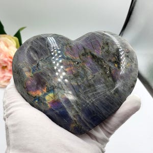 May include: A heart-shaped stone sculpture with a polished surface. The stone is primarily gray with iridescent flashes of purple, blue, and yellow. The sculpture is held in a white gloved hand.