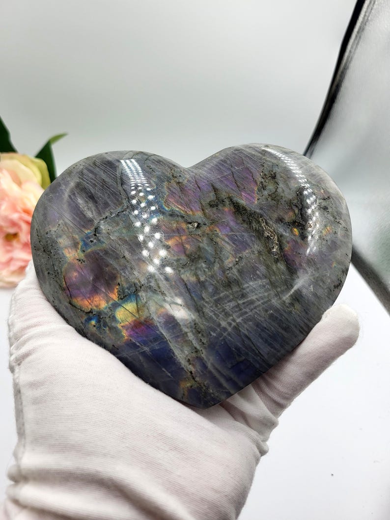 May include: A heart-shaped stone sculpture with a polished surface. The stone is primarily gray with iridescent flashes of purple, blue, and yellow. The sculpture is held in a white gloved hand.