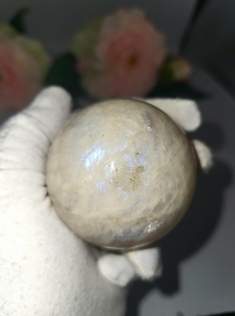 May include: A polished, spherical moonstone crystal. The stone is a creamy white color with iridescent blue and purple flashes. The sphere is held in a white gloved hand, with soft pink flowers in the background.