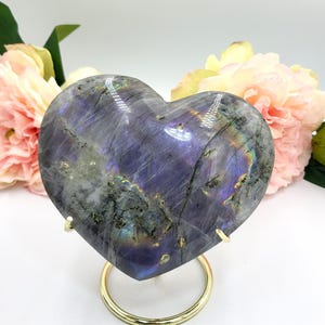 May include: A heart-shaped labradorite stone with iridescent blue, purple, and green hues. The polished stone is displayed on a gold-colored metal stand, with pink and yellow flowers in the background. A decorative item.