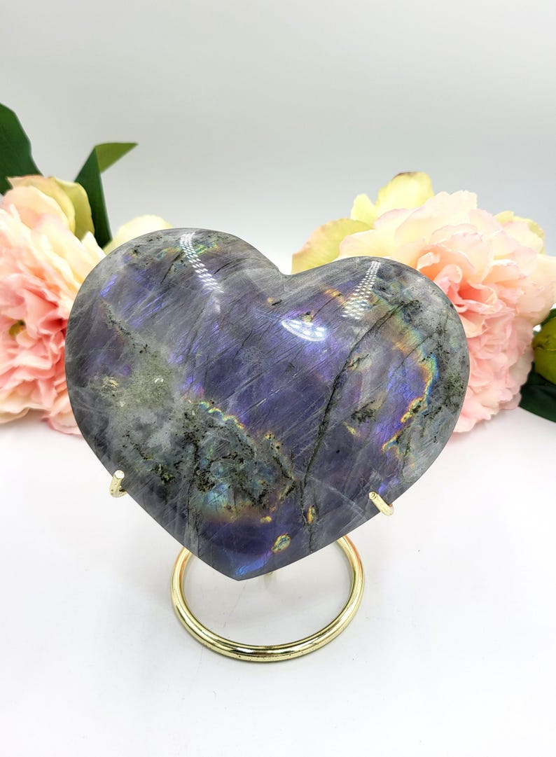 May include: A heart-shaped labradorite stone with iridescent blue, purple, and green hues. The polished stone is displayed on a gold-colored metal stand, with pink and yellow flowers in the background. A decorative item.