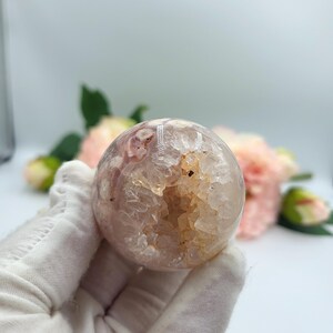 May include: A spherical, polished crystal geode with a hollow centre. The geode is primarily pink and white with areas of tan and clear crystal. The geode is held in a gloved hand, with pink flowers in the background.