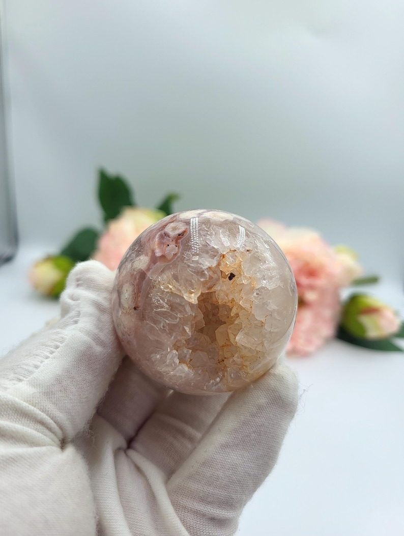 May include: A spherical, polished crystal geode with a hollow centre. The geode is primarily pink and white with areas of tan and clear crystal. The geode is held in a gloved hand, with pink flowers in the background.