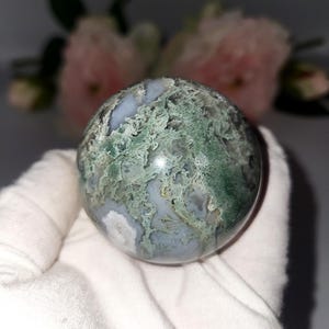 K&ouml;nnte beinhalten: Eine polierte Edelstein-Kugel mit einem Moosachat-Muster. Der Orb zeigt eine Mischung aus durchscheinenden wei&szlig;en, hellblauen und gr&uuml;nen Farbt&ouml;nen, die ein nat&uuml;rliches, organisches Aussehen erzeugen. Die Kugel wird in wei&szlig;em Stoff gehalten.