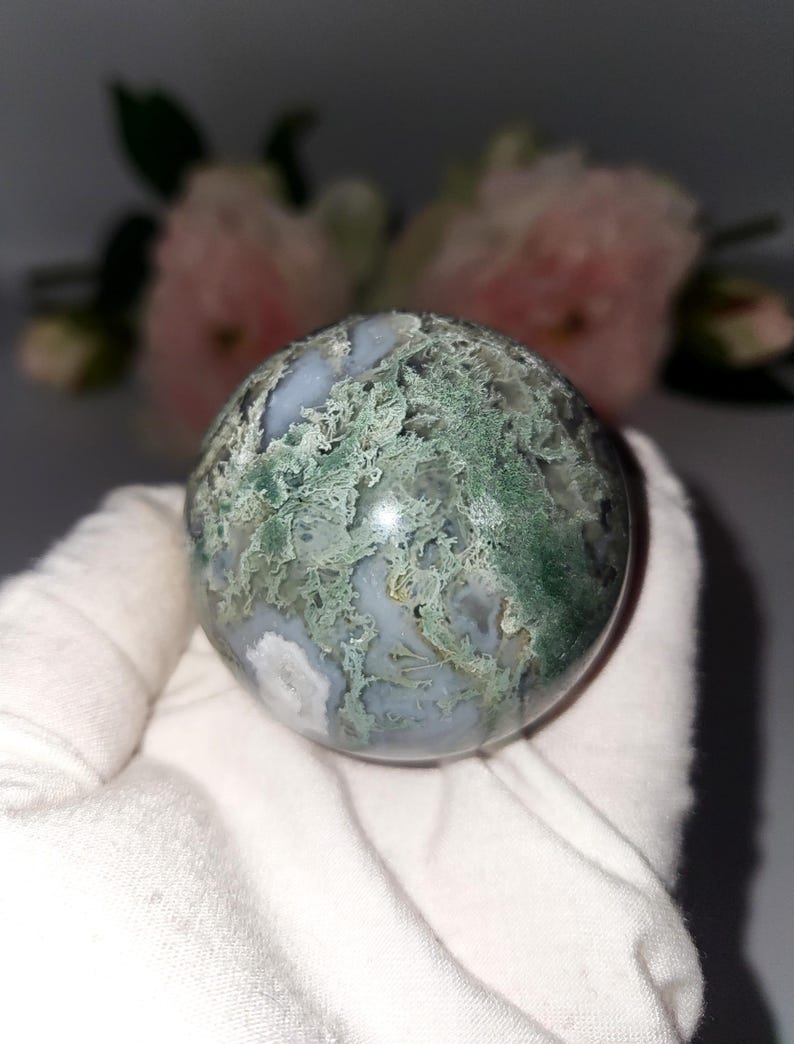 K&ouml;nnte beinhalten: Eine polierte Edelstein-Kugel mit einem Moosachat-Muster. Der Orb zeigt eine Mischung aus durchscheinenden wei&szlig;en, hellblauen und gr&uuml;nen Farbt&ouml;nen, die ein nat&uuml;rliches, organisches Aussehen erzeugen. Die Kugel wird in wei&szlig;em Stoff gehalten.
